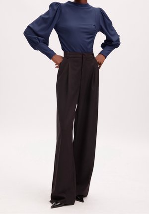 Vrouw draagt een marineblauwe blouse met pofmouwen, ingestopt in zwarte, wijde broek met hoge taille en zwarte puntige schoenen. Handen in de zij.