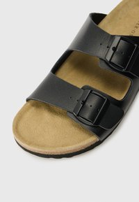 Sandala neagră din piele, tip șlapi, cu două curele reglabile și catarame negre, pe o talpă de culoare bej din piele întoarsă.