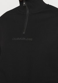 Sudadera negra con cremallera hecha de tela suave, con cuello alto, textura acanalada y "CALVIN KLEIN JEANS" bordado en hilo oscuro.
