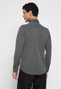 Grijze shirt met lange mouwen, een gestructureerde kraag en knoopmanchetten, gemaakt van gladde stof en met een gebogen zoomlijn. Gezicht naar achteren getoond.