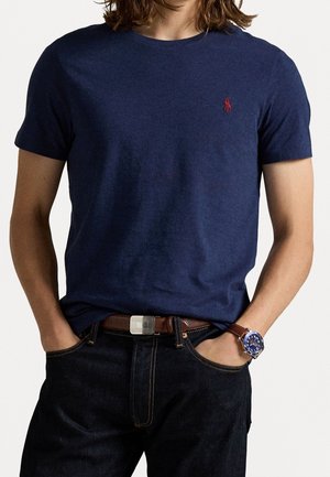 Mand iført en mørkeblå kortærmet t-shirt med rødt logo, mørke jeans, brunt bælte og blå ur med brun læderrem, hænderne i lommerne.