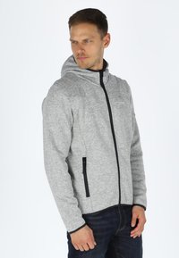 Felpa grigia con zip caratterizzata da un tessuto testurizzato, un cappuccio aderente e dettagli neri sulla zip, abbinata a jeans scuri. Design minimalista.