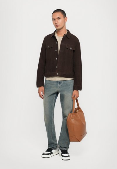 Veste en denim suédine marron avec un bouton à l'avant, chemise beiger, jeans bleu, baskets noir et blanc, tenant un sac tote en cuir marron.