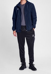 Granatowa lekka kurtka z kapturem, pod spodem szara koszulka, czarne sportowe spodnie z bocznymi paskami oraz czarne sportowe buty.