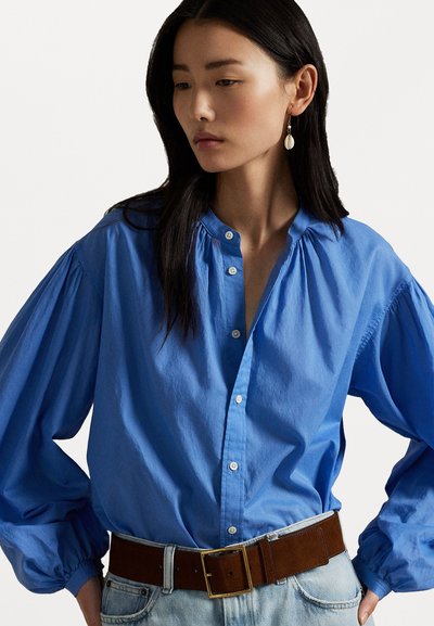 Blue cotton shirt with a buttoned front, pleated neckline, and puffed sleeves, paired with a brown leather belt and denim jeans.
