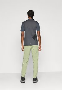 Γκρι πουκάμισο απομάκρυνσης υγρασίας με υφή υφάσματος και αναπνεύσιμη σχεδίαση, σε συνδυασμό με ανοιχτό πράσινο παντελόνι με ελαστικές μανσέτες και λεπτομέρεια με λογότυπο.