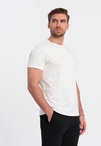 Hvid kortærmet t-shirt med et pletmønster, lavet af blødt stof; rund halsdesign, båret med sorte bukser.