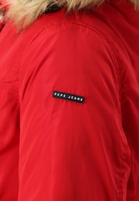 Piros puffertje, sima, fényes textúrával. A felső karon fekete "PEPE JEANS" logó található, és prémmel bélelt kapucni.