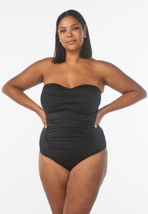 Selvsikker kvinde med medium hudtone og glat hår iført en sort, rynket, stropfri badedragt, hænderne på hofterne.