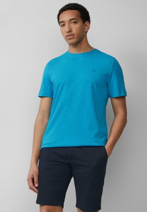 T-shirt bleu à manches courtes en tissu lisse, arborant un logo discret sur la poitrine. Associé à un short noir.