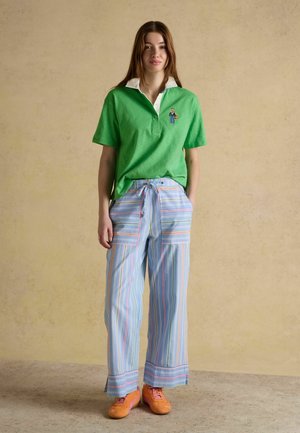 Jauna sieviete zaļā polo krekla, zilās svītrainās platajās biksēs un oranžās sporta apavos stāv pret bēša fona sienu.