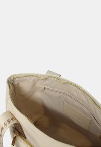 Interno della borsa a tracolla beige con tessuto liscio, tasca con zip e etichetta con logo, dotata di una cerniera color oro e texture morbida.