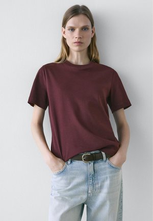 Vrouw met steil blond haar, gekleed in een bordeauxrood T-shirt met korte mouwen, ingestopt in een lichtblauwe spijkerbroek met een bruine riem, handen in de zakken.