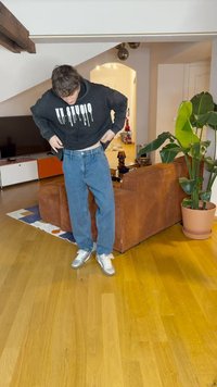 Melna kapuce ar baltu uzrakstu, kas pil, zilo denimā džinsos un sudraba kedās. Stāv uz koka grīdas blakus brūnai dīvānam un augiem.