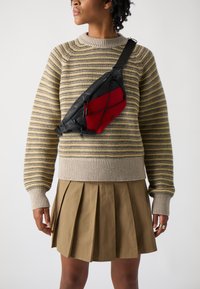 Schwarze und rote Umhängetasche mit geometrischem Design, ausgestattet mit einem strukturierten roten Paneel und elastischen Schnurdetaillierungen, getragen über einem gestreiften Pullover.