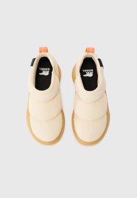 Zapatos slip-on beige con parte superior acolchada, suela texturizada color marrón, detalles en negro y tirador naranja. Logo visible en la plantilla.