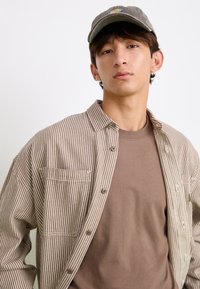 Brown and white striped shirt with a button-up design, paired with a plain brown t-shirt and a gray cap featuring a logo. Smooth fabric texture.