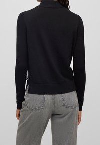 Un pull en tricot noir avec un col montant, un design ajusté, un ourlet côtelé et un détail de logo subtil. Assorti à un jean gris taille haute.