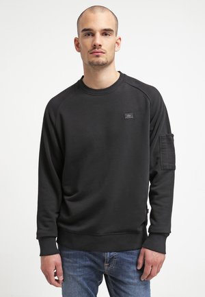 Sweatshirt noir avec col et poignets côtelés, présentant une poche plaquée sur la manche gauche. Fabriqué en matière polaire douce.