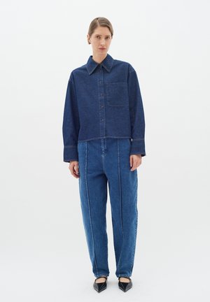 Dunkelblaue Jeanshemd, mit Knöpfen und Kragen, sowie einer aufgesetzten Tasche. Kombiniert mit hoch geschnittenen, locker sitzenden blauen Jeans mit sichtbaren Nähten.