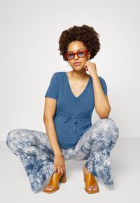 Top azul em V com cintura ajustável, combinado com calças largas em tie-dye azul e branco. Sandálias de salto abertas de cor castanha completam o look.