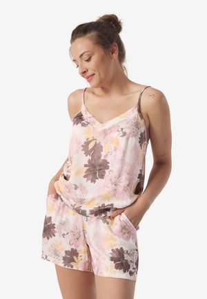 Kobieta z upiętymi włosami w koczek, ubrana w różowy, żółty i brązowy kwiecisty top na ramiączkach oraz dopasowane szorty, z rękami w kieszeniach, uśmiechnięta z zamkniętymi oczami.