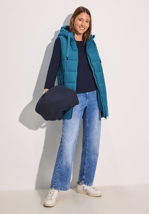 Tiefblaue Pufferweste mit Kapuze, navyblaues Langarmshirt, hellblaue weite Jeans und weiße Sneaker. Das Model hält ein navyblaues Kleidungsstück.