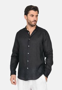 Camicia nera in lino con maniche lunghe e colletto button-down, dotata di bottoni bianchi e un taglio morbido, abbinata a pantaloni chiari.