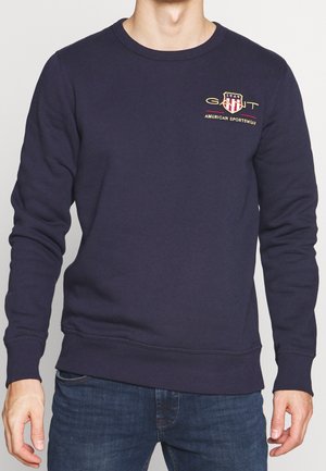 Mężczyzna ubrany w granatową bluzę z długim rękawem z logo GANT American Sportswear na górnej lewej części klatki piersiowej, w połączeniu z niebieskimi dżinsami.