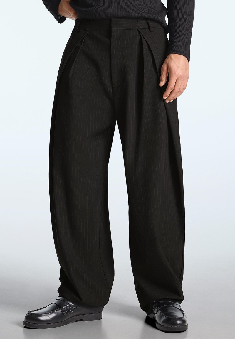 Muž nosící černé široké kalhoty s pruhy a černé kožené mokasíny, stojící s jednou rukou opřenou u kapsy.