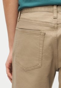 Άτομο που φοράει μπεζ παντελόνι με ορατή πίσω τσέπη και τυρκουάζ πουκάμισο που έχει βγει μέσα, δείχνοντας ραφές και θηλιές για ζώνη.