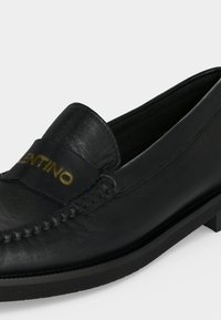 Mocassin en cuir noir avec des détails cousus et un logo doré "VALENTINO" sur le dessus, doté d'un petit talon superposé et d'un bout arrondi.