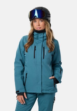Une femme porte une veste et un pantalon de ski bleu sarcelle, un casque noir et des lunettes de ski à verres réfléchissants, sur un fond uni.
