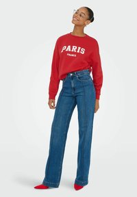 Rode sweatshirt met "PARIS FRANCE" in witte tekst, gecombineerd met hooggesneden, wijde blauwe jeans. Heeft rode puntige schoenen.