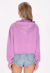 Maglione all'uncinetto viola con cappuccio regolabile, design cropped e maniche lunghe. Abbinato a pantaloni shorts di jeans, che evidenziano una vestibilità rilassata.