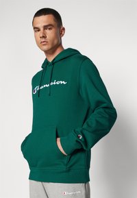 Groene capuchontrui met een voorktas, met een wit "Champion" logo. Geribde manchetten en zoom, zachte textuur, losse pasvorm.