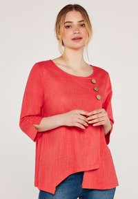 Korallfarbenes Leinenblusenshirt mit drei Holzknöpfen an der linken Schulter, runder Ausschnitt und drei Viertelärmeln. Asymmetrischer Saum.