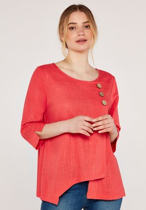 Korallfarbenes Leinenblusenshirt mit drei Holzknöpfen an der linken Schulter, runder Ausschnitt und drei Viertelärmeln. Asymmetrischer Saum.