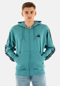 Giovane uomo che indossa una felpa con zip color teal e strisce nere sulle maniche, in piedi con le mani nelle tasche frontali su uno sfondo bianco.