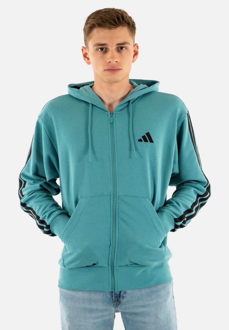 Giovane uomo che indossa una felpa con zip color teal e strisce nere sulle maniche, in piedi con le mani nelle tasche frontali su uno sfondo bianco.