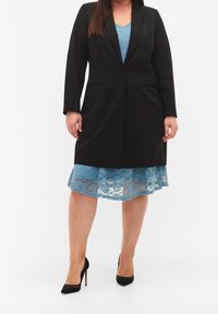 Blazer noir à manches longues, avec deux poches et une coupe ajustée, sur une robe bleue en dentelle avec un ourlet festonné. Escarpins noirs.