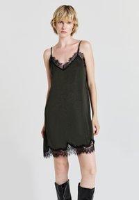 Donna con capelli biondi corti che indossa un abito nero a sottoveste con pizzo e stivali neri fino al ginocchio, su uno sfondo chiaro e semplice.