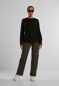 Maglione nero a costine, pantaloni cargo verde oliva con tasche e cerniere, e sneakers bianche. Gli occhiali da sole aggiungono contrasto all'outfit.