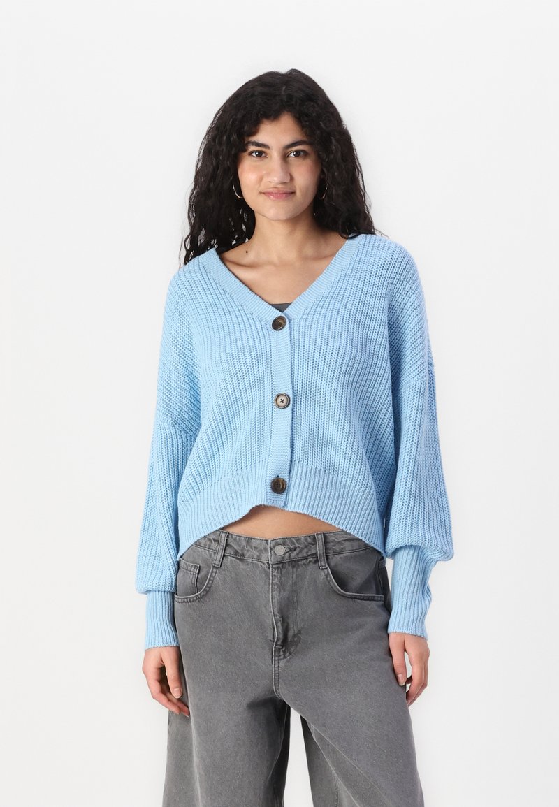 lichtblauwe cropped cardigan met een V-hals, geribde textuur en drie grote knopen aan de voorkant. Heeft losse mouwen en een boxy ontwerp.