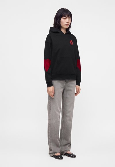 Femme portant un sweat à capuche noir avec des patchs en forme de cœur rouge aux coudes, un jean gris à jambes larges, et des ballerines noires, debout devant un fond blanc uni.