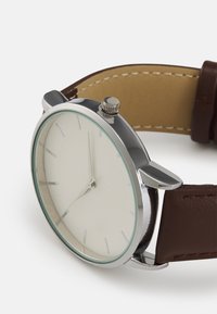 Montre ronde en argent avec un cadran blanc, index d'heure minimalistes et un bracelet en cuir marron. Présente une couronne texturée et des accents de surpiqûres légères.