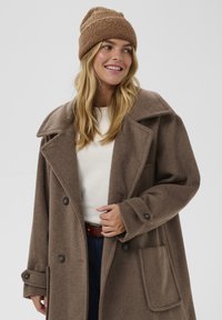 Palton maro supradimensionat cu rever mari și închidere cu nasturi dubli, asociat cu o ținută dintr-o căciulă maro texturată. Cămașă albă dedesubt.