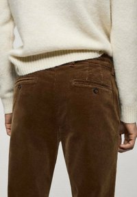 Pantalon en velours côtelé marron avec des côtes verticales, doté de deux poches arrière et de petits accents de boutons, associé à un pull crème.