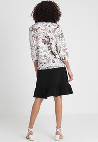 Weißes Oberteil mit floralen Mustern und Dreiviertelärmeln, kombiniert mit einem schwarzen Rock mit Rüschen. Absatzschuhe mit Knöchelriemen vollenden den Look.