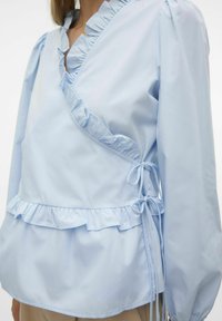 Blusa azzurro chiaro con scollatura a V, rifiniture arruffate e dettagli con coulisse ai lati, maniche gather e orlo a peplum.